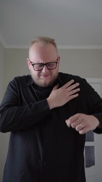 Middle-aged man in glasses grasps his shoulder and winces, reacting to sudden upper body pain in a neutral indoor setting.