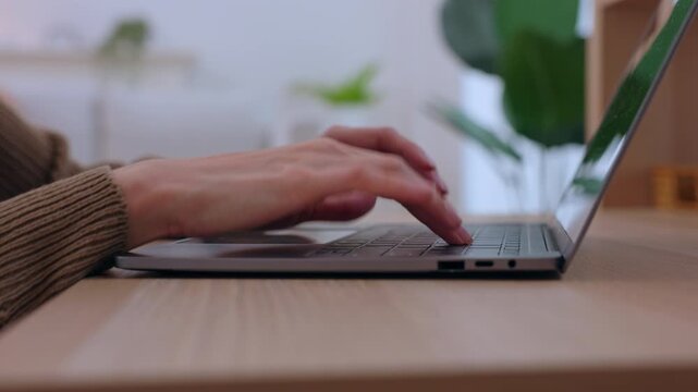 Laptop hand typing on keyboard at wooden desk computer work from home office with plant close view remote online workspace soft light