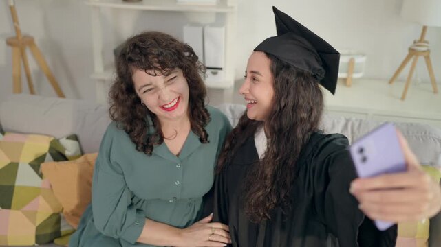 Young Latino daughter wearing graduation gowns taking selfies with mother. 