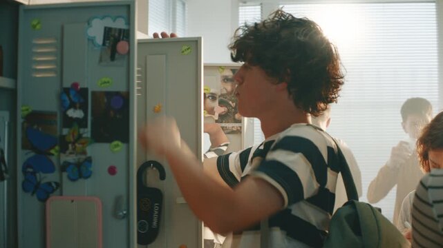 Teenage Schoolboy Searches for Lost Key to Locker in High School Hallway During Class Break, Reaching Among Lockers Classmates were Gathered Around. Authentic Student Life and Everyday School Routine.