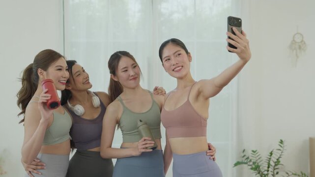 Young Asian women fitness friends taking smartphone selfie after exercise bright studio with water bottle and headphones, smiling together friendship and healthy lifestyle celebration indoors with