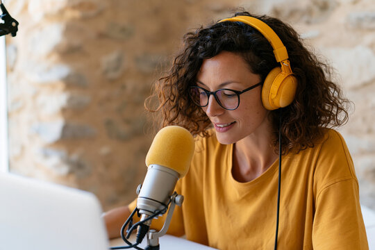 Woman smiles while recording podcast or voiceover. She wears yellow headphones and glasses. Her curly hair frames her focused expression. Soft lighting enhances cozy, creative atmosphere