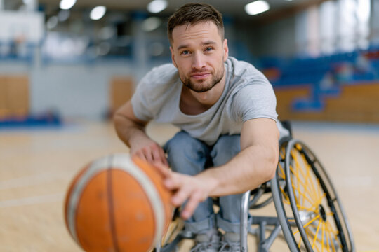 Young man smiles while seated in a wheelchair. He grips a basketball, ready to play. The gymnasium floor reflects soft, natural light. He looks directly at the viewer with confidence