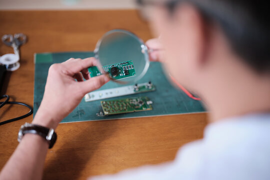 Mature Asian man looks at and considers circuit board details with magnifying glass, focusing on precision checks for home electrical appliance DIY fix, electronics component hobby, handyman workshop.