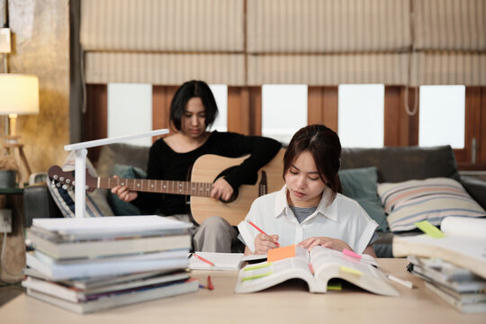 Two Asian teenage tutorials, girl concentrates on reading books for exam, learning at home, her friend relaxes by playing guitar music, self-study with textbooks, homeschooling together lifestyle.