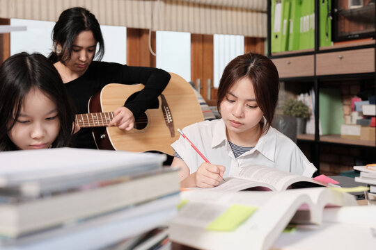 Three Asian teenage tutorials, girls concentrate on reading books for exams, learning at home, her friend relaxes by playing guitar music, self-study with textbooks, homeschooling together lifestyle.