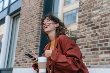 Fototapeta premium Young woman laughing while holding a phone and coffee cup on a city street, featuring warm earthy tones for lifestyle and telecom marketing