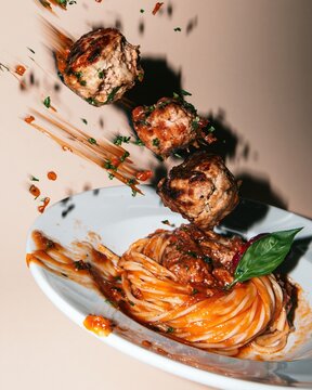 Meatballs falling toward a plate of spaghetti with glossy tomato sauce and fresh basil, mid-air for food packaging and delivery ads