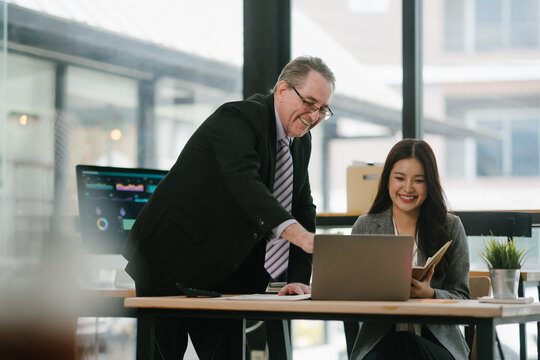 Experienced manager mentoring young professional, discussing project data on a laptop in a contemporary coworking space, smiling