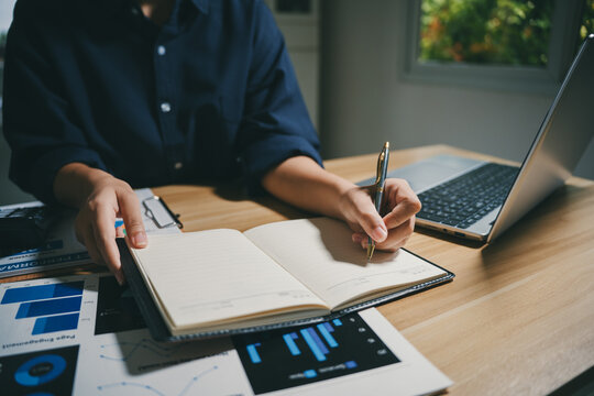 Person hands writing in a notebook, working on business data with charts and a laptop on a wooden desk