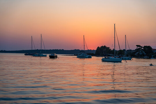 Sailing yachts anchored in a calm bay during a colorful summer sunset over the sea. Peaceful evening, golden hour, coastal travel destination and relaxing sailing lifestyle.