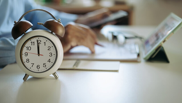 close up shot, focus alarm clock 12.00 PM with blurred businessman working with tablet and business document before breaking lunch
