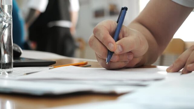 Close-up of a person writing on paperwork at a desk. Filling out documents, application forms or registration papers in an office or administrative setting.