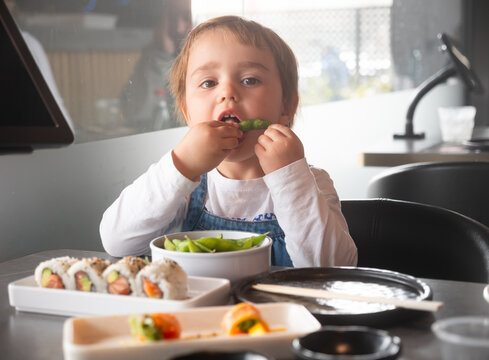 Child sits on a high chair near a table and eats edamame beans. Child tastes beans and rolls