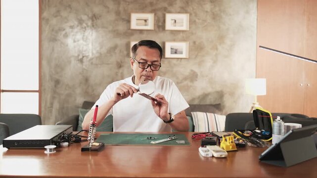 Mature Asian man looks at and considers circuit board details with magnifying glass, focusing on precision checks for home electrical appliance DIY fix, electronics component hobby, handyman workshop.