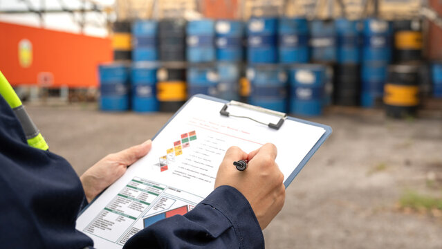 Checking on chemical hazardous safety checklist form, close-up at worker hand, with factory warehouse and metal barrel storage as background. Industrial safe work practice scene, selective focus.