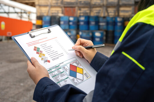 Checking on chemical hazardous safety checklist form, close-up at worker hand, with factory warehouse and metal barrel storage as background. Industrial safe work practice scene, selective focus.