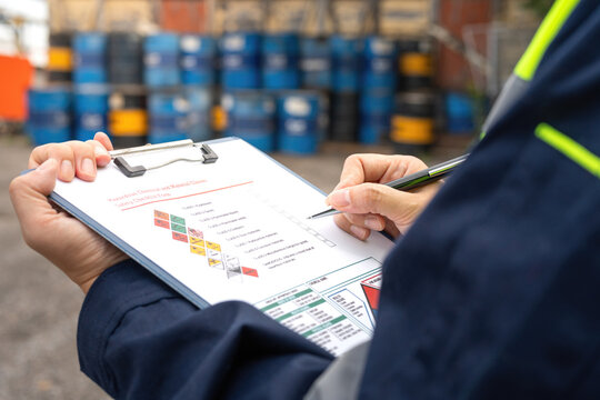 Checking on chemical hazardous safety checklist form, close-up at worker hand, with factory warehouse and metal barrel storage as background. Industrial safe work practice scene, selective focus.