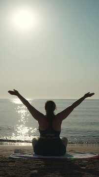 Silhouette of young woman practicing yoga on seashore in lotus pose, vertical video. Female figure raising arms sideways up and lowering to chest for breathing exercise.