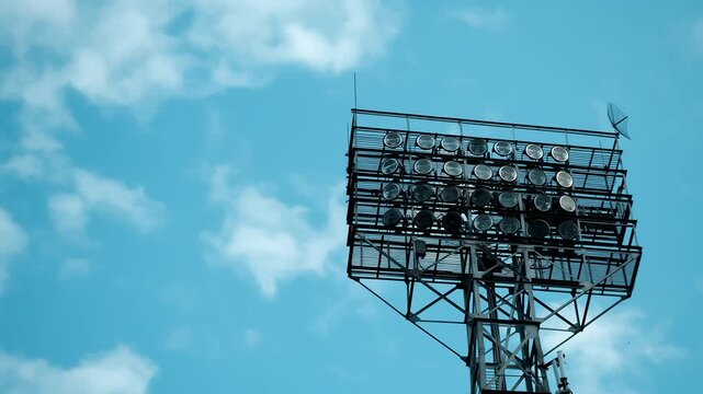 Large array of stadium floodlights mounted on a tall metal tower against blue sky. Powerful outdoor lighting equipment for sports arenas and large venues.