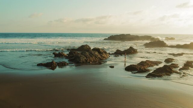 Slender woman walking along rocky beach shore at sunset with ocean waves and golden light