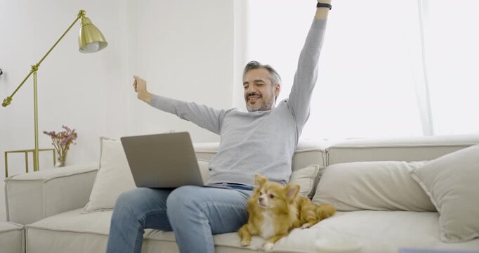 Cheerful caucasian senior man sitting on a comfortable sofa with laptop, raising his arms in celebration during a successful work stretching leisure moment.