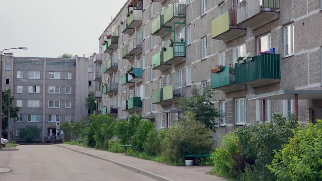 Handheld tracking shot along sidewalk near multi story panel apartment building with colorful balconies and green bushes in Daugavpils suburban neighborhood.
