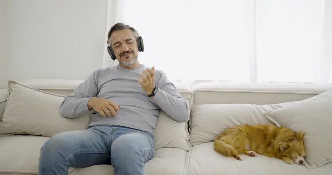 Portrait of senior old caucasian man stay home sit on the sofa  living room listen to fun music head phone with lovely dog. Happy moment of pure love middle age male playing with little dog in winter