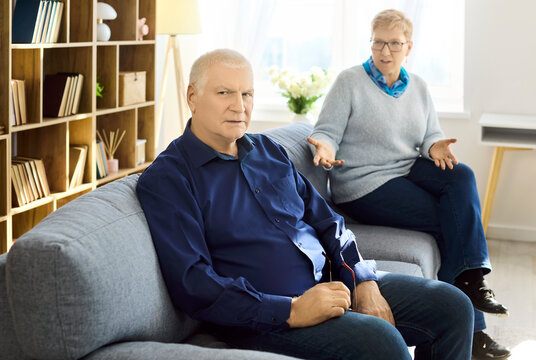 Senior man and woman couple argument conflict stress. Older husband on sofa looks upset as wife gestures, showing frustration and relationship problems at home. Clear theme of couple conflict.