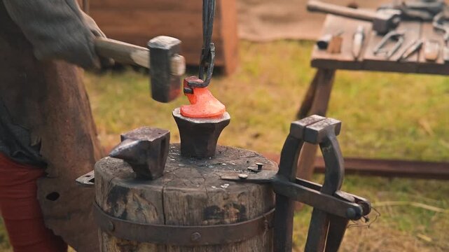 A blacksmith shapes heated metal using a hammer on an anvil in a medieval setting. The person is wearing simple, period-appropriate clothing and is focused on the task