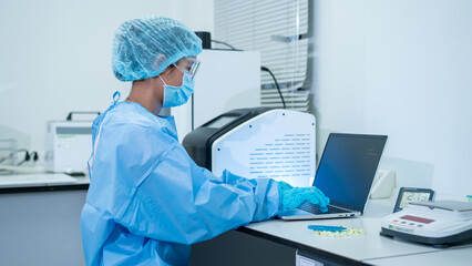 A laboratory worker wearing protective gear uses a laptop in a clean medical lab with equipment and test supplies. © NanSan