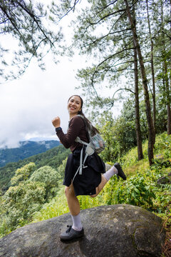 And keywordsSmiling Asian female hiker wearing backpack and standing on forest boulder with mountain landscape, pine tree, and lush vegetation during outdoor adventure