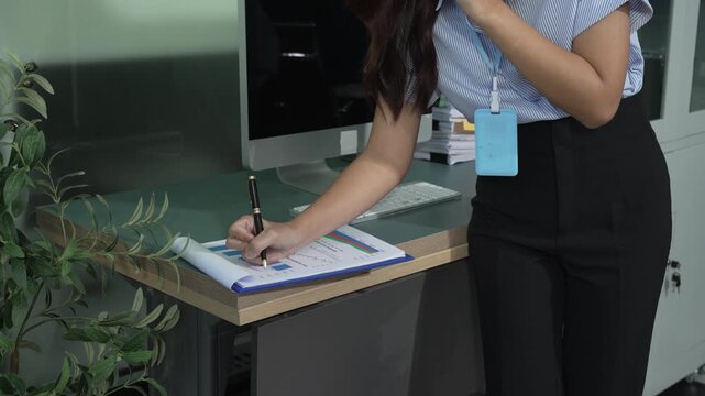 Businesswoman writing on a clipboard beside an office desk and computer, with ID badge, indoor workplace scene showing planning, paperwork and professional routine.