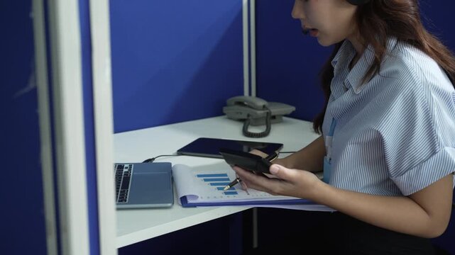 Woman in a small office booth using a smartphone while working with a laptop and paperwork, wearing a headset for customer support or remote business communication.