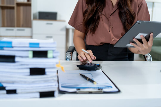Businesswoman at office desk using a tablet and calculator beside stacks of paperwork, showing accounting, budgeting, finance, and busy administrative work.