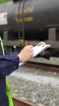 A safety officer is checking on hazardous chemical material checklist for risk assessment before tranportation the dangerous good with train tanker railway background. Industrail working scene.