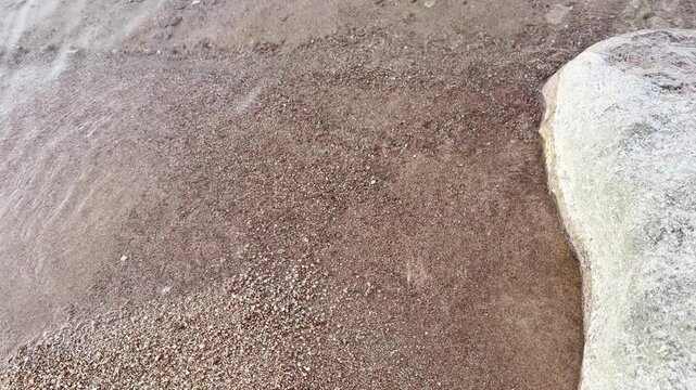 Natural pink sandy shoreline with clear shallow water, gentle ripples and a pale textured rock. Soft coastal colors and organic textures create a serene summer nature background for travel and design