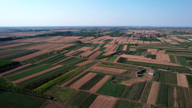 Aerial shot of patchwork farmlands with varied crops and soil under a clear blue sky, showing rural agricultural landscape.