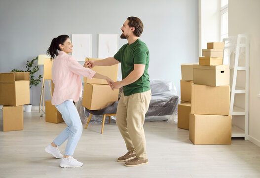 Couple dancing while moving boxes in home. Smiling man and woman enjoy happiness while unpacking in a bright room with stacked cardboard boxes. Relocation success, love, and teamwork theme.