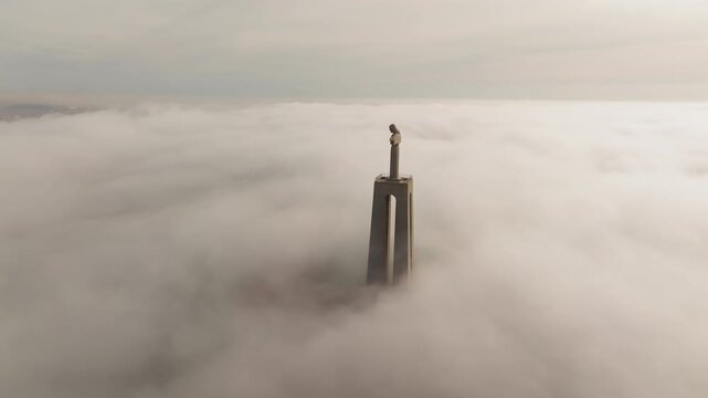 Cristo Rei statue, Almada, Lisbon. Slow orbit drone shot at sunrise. Dense fog blankets city. Statue dramatically silhouetted above horizon