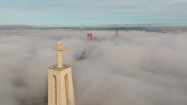 Drone flies toward Cristo Rei statue above dense fog, Lisbon, Almada. 25 de Abril Bridge top emerges from mist at sunrise. Cinematic Portugal aerial footage