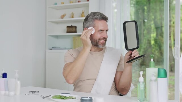 Man doing male skincare routine at home. Mature man applying cosmetic product on face. Male grooming and skincare routine concept. Bearded man using facial toner with cotton pad.