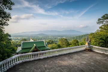Fototapeta premium Green roof temple viewpoint in Khao Yai, Thailand, offers wide panorama of lush green forest valley and distant blue mountains. Stone statue decorates white railing on terrace.