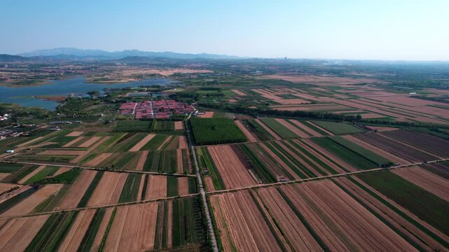 This aerial shot shows patchwork farmland, a reservoir, and small settlements under a clear blue sky with distant mountains.