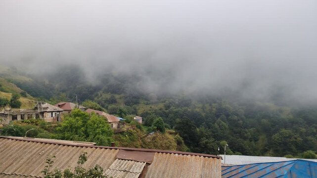Thick fog and clouds rolling over a mountain village and forest. Beautiful view of the mountains in the morning. Fog covered the houses of the village. Green forests and fields and white fog.