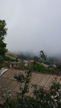 Thick fog and clouds rolling over a mountain village and forest. Beautiful view of the mountains in the morning. Fog covered the houses of the village. Green forests and fields and white fog.