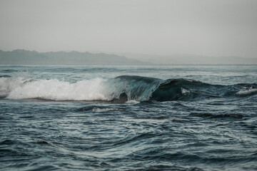 Naklejka premium Misty seascape of rolling teal waves breaking along a distant hazy coastline under an overcast gray sky