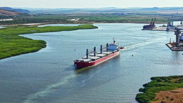 A cargo ship navigates a river toward a port where loading and unloading activities take place. Green fields and hills surround the area during the day.