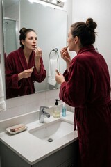 Obraz premium Young woman in red bathrobe applies lip balm in bathroom mirror. Female person uses cosmetic lip care product for skin hydration. Morning beauty routine at home with wellness skincare habit for soft