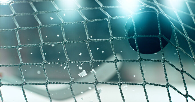 Action sports photo of a hockey puck flying into the goal net during a scoring shot, with ice particles in the air and a hockey stick visible in the background, capturing speed and intensity.